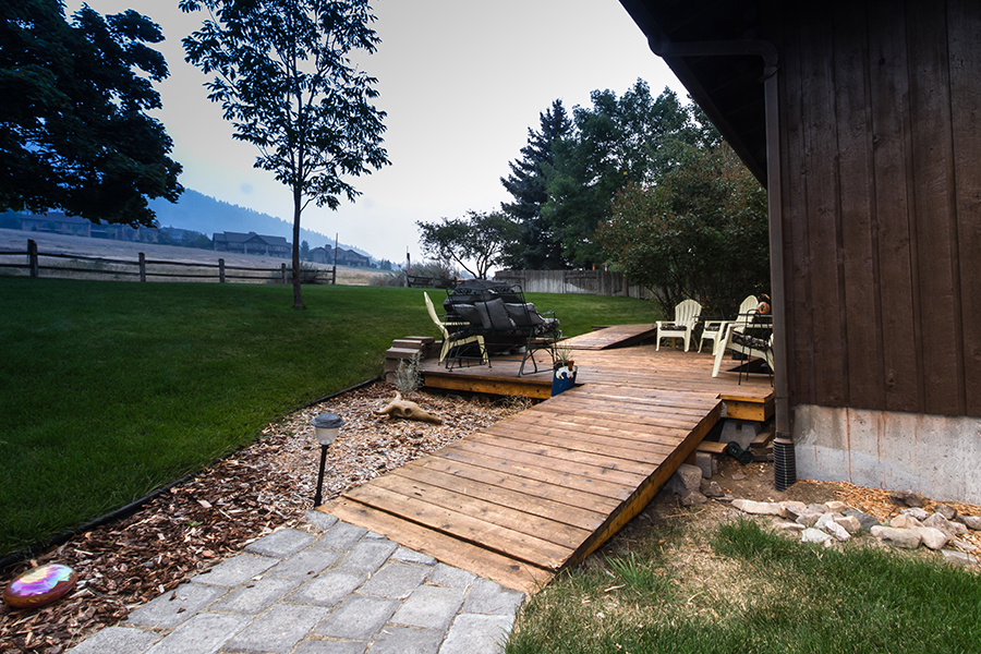 A short wooden ramp provides a path of travel up onto a low deck covered with lawn chairs, a grill and an outdoor loveseat. In the background, another short wooden ramp goes up to the next level of the deck. A decorative shiny rainbow-hued stone and bleached cow skull sit in the garden near the deck.